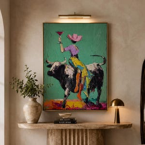Peut inclure: Une peinture à l'huile encadrée représentant une silhouette portant un chapeau de cow-boy rose et une chemise violette, montée sur un taureau noir et blanc tout en tenant un verre à cocktail rouge, avec des coups de pinceau texturés sur fond vert.