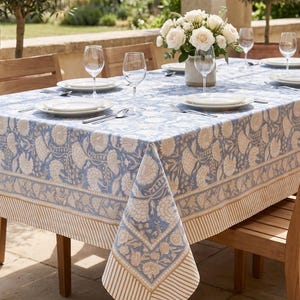 Peut inclure: Une nappe rectangulaire ornée d'un motif floral bleu clair et blanc avec une bordure rayée. Le tissu recouvre une table en bois dressée avec des assiettes blanches, des verres transparents et un vase central de roses blanches.