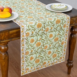 Può includere: Un runner da tavola rettangolare con sfondo verde chiaro e un motivo ripetuto di fiori arancioni e viticci verde scuro. Il runner presenta un bordo decorativo ed è posato su un tavolo in legno con piatti di frutta.