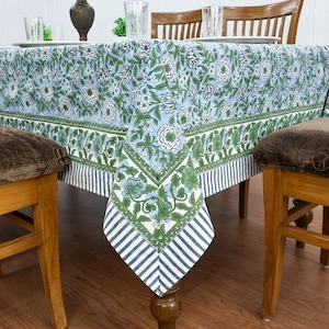 Puede incluir: Un mantel floral azul claro con detalles en verde y blanco. El mantel presenta un patrón repetido de flores y hojas. El borde tiene un diseño floral verde y un borde a rayas. El mantel está sobre una mesa de madera.