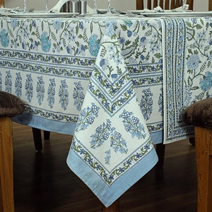 Può includere: Una tovaglia bianca con un motivo floreale blu e verde. La tovaglia ha un bordo con un motivo ripetitivo di foglie e fiori. La tovaglia è stesa su un tavolo di legno.