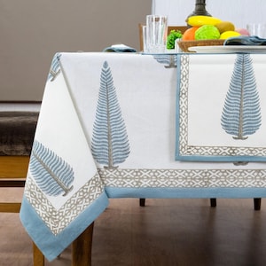 Puede incluir: Mantel blanco con diseño de árbol azul y gris. El mantel tiene un borde azul y un patrón geométrico gris. También se ven servilletas con el mismo diseño de árbol.