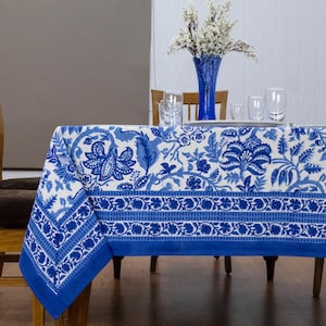 May include: A white and blue floral tablecloth covers a wooden table. The tablecloth features a repeating pattern of blue flowers and leaves on a white background. A blue border frames the edge. A blue vase with white flowers and glasses are on the table.