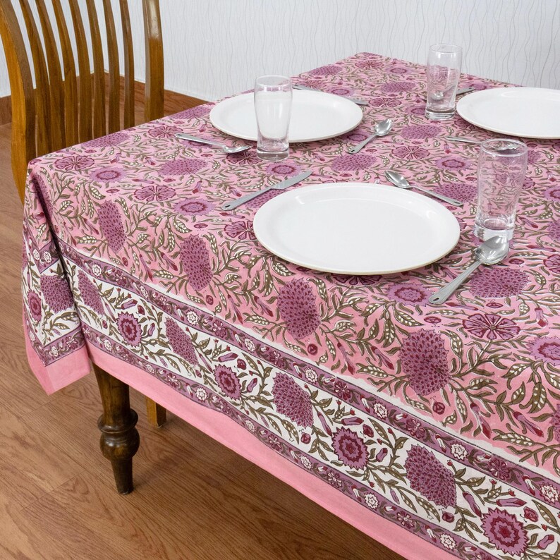 Beautiful hand block printed cotton tablecloth featuring floral patterns with pink and red accents. This vibrant Indian dining table cover adds a touch of bohemian charm to your kitchen or dining space. Ideal for everyday use, festive meals, or special occasions like housewarmings, weddings, baby showers, or dinner parties. Styled on a wooden table with white plates, cutlery, glasses, and a centerpiece flower vase.