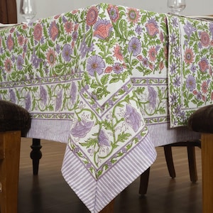 Puede incluir: Un mantel floral blanco con un patrón repetido de flores y hojas rosas, moradas y verdes. El mantel tiene un borde a rayas de color púrpura claro. La mesa está puesta con vasos y platos.