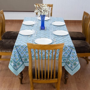 Puede incluir: Una mesa de comedor para seis personas con un mantel azul claro con estampado floral. Platos blancos, vasos transparentes y cubiertos están dispuestos alrededor de un jarrón azul con flores blancas. Sillas de madera con asientos acolchados marrones rodean la mesa.