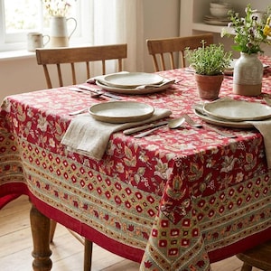 Pode incluir: Uma toalha de mesa retangular com base em vermelho profundo, padrão floral multicolorido e borda geométrica. A mesa está posta com pratos de cerâmica, guardanapos de linho, talheres de metal, uma planta verde e um vaso de cerâmica.