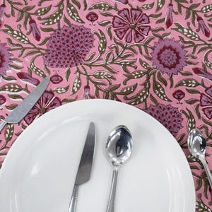 Beautiful hand block printed cotton tablecloth featuring floral patterns with pink and red accents. This vibrant Indian dining table cover adds a touch of bohemian charm to your kitchen or dining space. Ideal for everyday use, festive meals, or special occasions like housewarmings, weddings, baby showers, or dinner parties. Styled on a wooden table with white plates, cutlery, glasses, and a centerpiece flower vase.