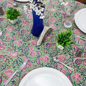 Beautiful hand block printed cotton tablecloth featuring floral patterns with pink and green accents. This vibrant Indian dining table cover adds a touch of bohemian charm to your kitchen or dining space. Ideal for everyday use, festive meals, or special occasions like housewarmings, weddings, baby showers, or dinner parties. Styled on a wooden table with white plates, cutlery, glasses, and a centerpiece flower vase.
