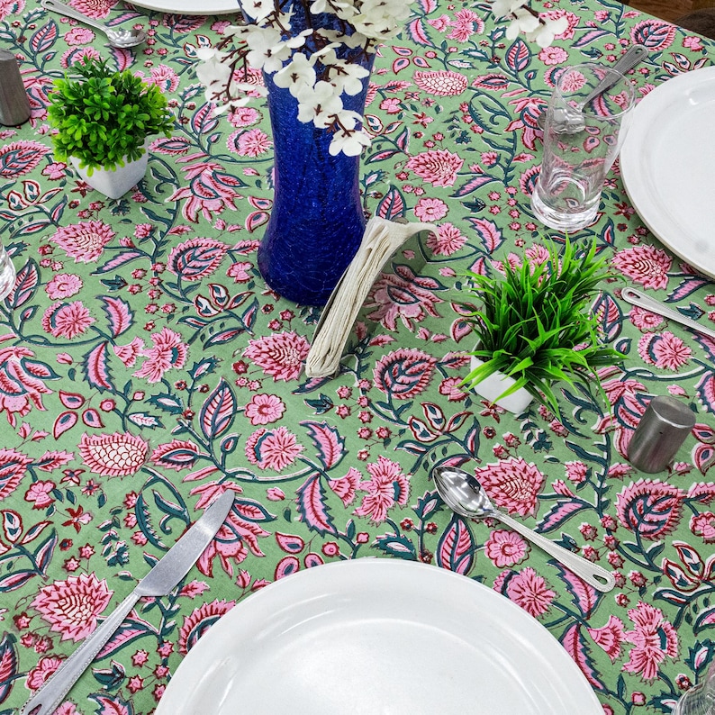 Beautiful hand block printed cotton tablecloth featuring floral patterns with pink and green accents. This vibrant Indian dining table cover adds a touch of bohemian charm to your kitchen or dining space. Ideal for everyday use, festive meals, or special occasions like housewarmings, weddings, baby showers, or dinner parties. Styled on a wooden table with white plates, cutlery, glasses, and a centerpiece flower vase.