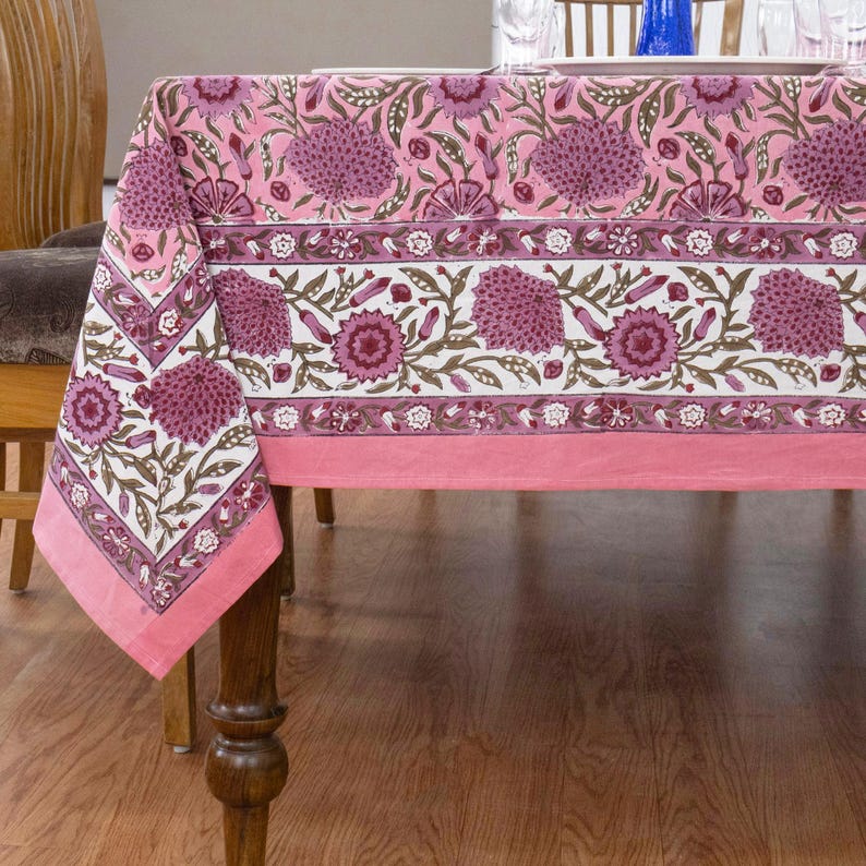 Beautiful hand block printed cotton tablecloth featuring floral patterns with pink and red accents. This vibrant Indian dining table cover adds a touch of bohemian charm to your kitchen or dining space. Ideal for everyday use, festive meals, or special occasions like housewarmings, weddings, baby showers, or dinner parties. Styled on a wooden table with white plates, cutlery, glasses, and a centerpiece flower vase.