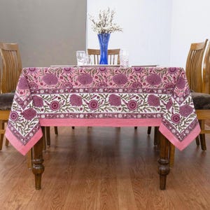 Beautiful hand block printed cotton tablecloth featuring floral patterns with pink and red accents. This vibrant Indian dining table cover adds a touch of bohemian charm to your kitchen or dining space. Ideal for everyday use, festive meals, or special occasions like housewarmings, weddings, baby showers, or dinner parties. Styled on a wooden table with white plates, cutlery, glasses, and a centerpiece flower vase.