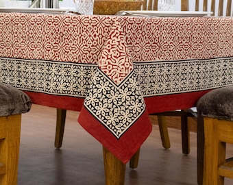 May include: A rectangular tablecloth featuring a repeating geometric floral pattern in deep red, black, and cream. The fabric has a wide border with a distinct black and cream floral motif, finished with a solid red edge draped over a wooden table.