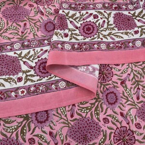 Beautiful hand block printed cotton tablecloth featuring floral patterns with pink and red accents. This vibrant Indian dining table cover adds a touch of bohemian charm to your kitchen or dining space. Ideal for everyday use, festive meals, or special occasions like housewarmings, weddings, baby showers, or dinner parties. Styled on a wooden table with white plates, cutlery, glasses, and a centerpiece flower vase.