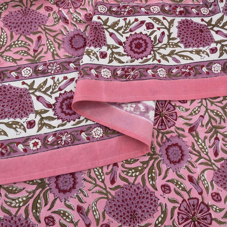 Beautiful hand block printed cotton tablecloth featuring floral patterns with pink and red accents. This vibrant Indian dining table cover adds a touch of bohemian charm to your kitchen or dining space. Ideal for everyday use, festive meals, or special occasions like housewarmings, weddings, baby showers, or dinner parties. Styled on a wooden table with white plates, cutlery, glasses, and a centerpiece flower vase.