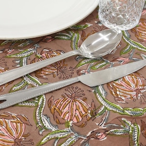 Puede incluir: Primer plano de una mesa con un plato blanco, un vaso transparente y cubiertos. Los cubiertos incluyen una cuchara y un cuchillo. La mesa está cubierta con un mantel marrón con estampado floral con acentos rosas y verdes.