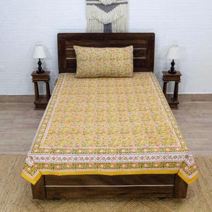 Puede incluir: Un juego de cama con estampado floral amarillo con una almohada a juego. El juego de cama presenta un patrón repetido de flores rosas y hojas verdes sobre un fondo amarillo. La cama está hecha y lista para usar.