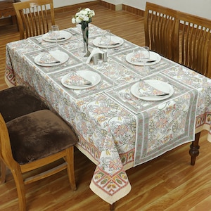 Puede incluir: Un mantel blanco con un patrón floral y un borde de rojo, naranja y verde. El mantel está puesto con seis cubiertos, cada uno con un plato blanco, una servilleta blanca y un vaso. La mesa es de madera y tiene un acabado marrón.
