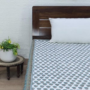 May include: A white bed sheet with a repeating pattern of blue floral designs. The sheet is on a wooden bed frame with a white pillow. A decorative border with a blue and brown pattern runs along the edge of the sheet.