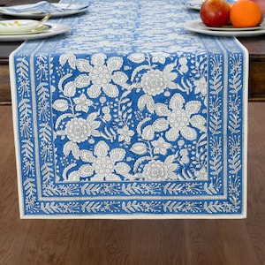May include: A rectangular table runner featuring a vibrant blue background with a white floral pattern. The design includes large, detailed blossoms and leaves in the center, framed by a decorative border of smaller leaves and vines.