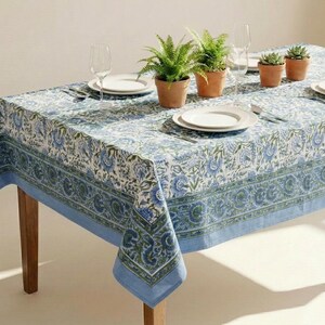 May include: A rectangular tablecloth featuring a white base with a repeating blue and green floral block print pattern, finished with a wide decorative border. The table is set with white plates, clear glasses, and three small potted green plants.