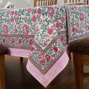 May include: A white tablecloth with a pink and green floral print. The tablecloth has a pink and white striped border. The tablecloth is draped over a table and chairs.