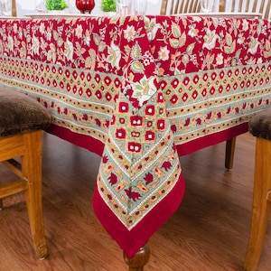 Peut inclure: Une nappe rouge ornée de motifs floraux et d'une bordure décorative. La nappe est drapée sur une table en bois, mettant en valeur son design. La bordure présente un motif géométrique avec des accents rouges, verts et dorés. Thème : décoration intérieure.