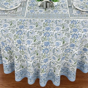 May include: A white tablecloth featuring a repeating floral pattern in shades of blue and green. The design includes stylized flowers and foliage with a solid light blue border along the bottom edge. The fabric drapes over a table surface.