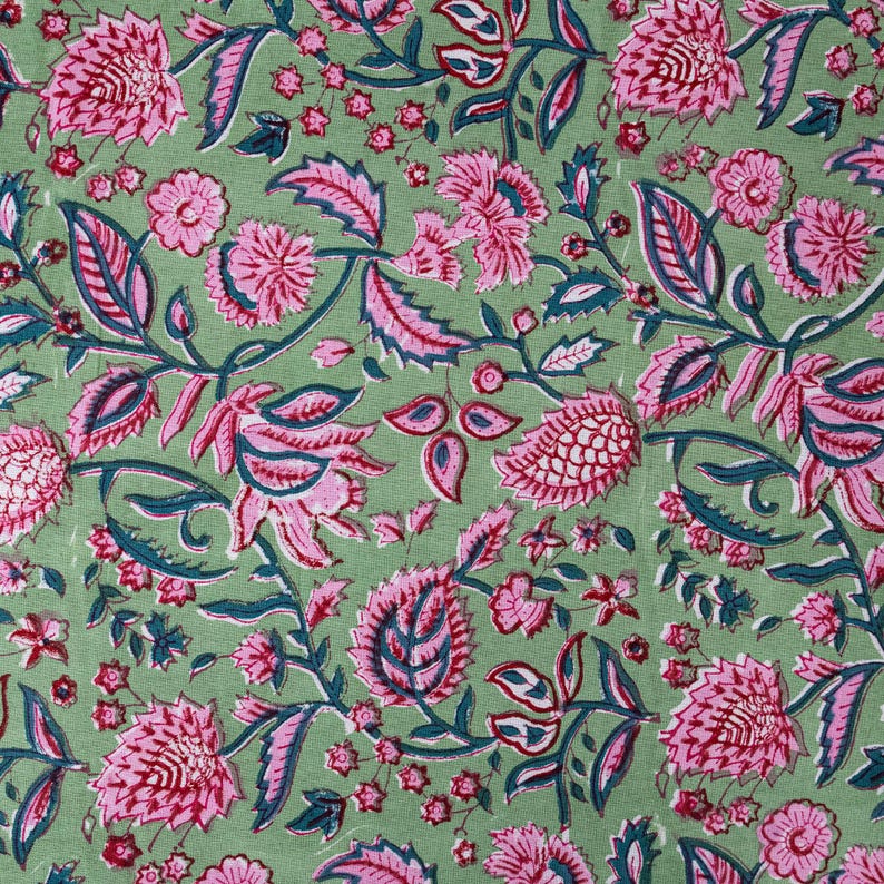 Beautiful hand block printed cotton tablecloth featuring floral patterns with pink and green accents. This vibrant Indian dining table cover adds a touch of bohemian charm to your kitchen or dining space. Ideal for everyday use, festive meals, or special occasions like housewarmings, weddings, baby showers, or dinner parties. Styled on a wooden table with white plates, cutlery, glasses, and a centerpiece flower vase.