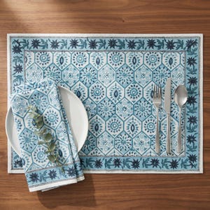 May include: A rectangular placemat and matching folded napkin featuring a light blue and white geometric hexagonal pattern with a dark blue floral border. A white ceramic plate, a silver fork, a silver knife, and a silver spoon are included.