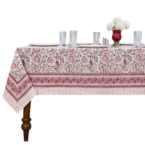 May include: A rectangular tablecloth featuring a repeating floral pattern in shades of pink and brown on a white background. The fabric has a wide border with a smaller floral motif and a geometric band at the bottom edge, draped over a wooden table.