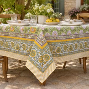 May include: A square tablecloth featuring a block-printed floral pattern in shades of yellow, green, and lavender. The design includes a central botanical motif, a wide decorative border with yellow and green flowers, and a striped yellow hem.