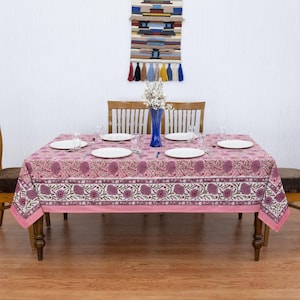 Beautiful hand block printed cotton tablecloth featuring floral patterns with pink and red accents. This vibrant Indian dining table cover adds a touch of bohemian charm to your kitchen or dining space. Ideal for everyday use, festive meals, or special occasions like housewarmings, weddings, baby showers, or dinner parties. Styled on a wooden table with white plates, cutlery, glasses, and a centerpiece flower vase.