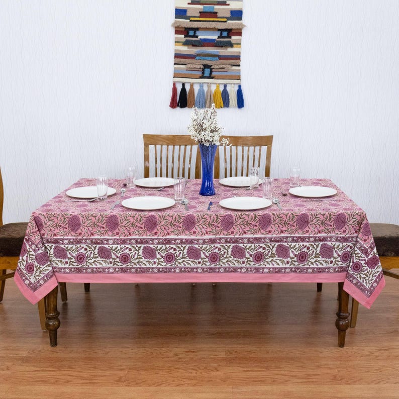 Beautiful hand block printed cotton tablecloth featuring floral patterns with pink and red accents. This vibrant Indian dining table cover adds a touch of bohemian charm to your kitchen or dining space. Ideal for everyday use, festive meals, or special occasions like housewarmings, weddings, baby showers, or dinner parties. Styled on a wooden table with white plates, cutlery, glasses, and a centerpiece flower vase.
