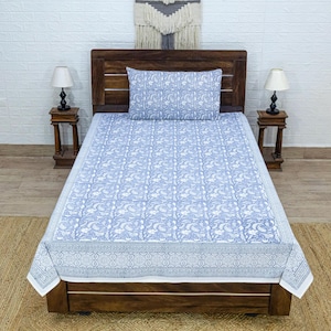 May include: A blue and white patterned bedspread and pillow set on a wooden bed frame. The bedding features a repeating floral design. Two small wooden side tables with lamps flank the bed. A woven wall hanging is above the bed.