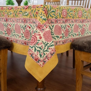 Könnte beinhalten: Ein gelbes Tischtuch mit Blumenmuster und einem rosa-grünen Blumenmuster. Das Tischtuch ist über einem Holztisch drapiert. Das Tischtuch hat einen Rand aus rosa und grünen Blumendesigns. Die Ecke des Tischtuchs hängt herunter.