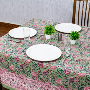 Beautiful hand block printed cotton tablecloth featuring floral patterns with pink and green accents. This vibrant Indian dining table cover adds a touch of bohemian charm to your kitchen or dining space. Ideal for everyday use, festive meals, or special occasions like housewarmings, weddings, baby showers, or dinner parties. Styled on a wooden table with white plates, cutlery, glasses, and a centerpiece flower vase.