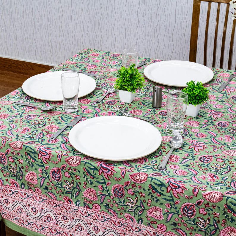 Beautiful hand block printed cotton tablecloth featuring floral patterns with pink and green accents. This vibrant Indian dining table cover adds a touch of bohemian charm to your kitchen or dining space. Ideal for everyday use, festive meals, or special occasions like housewarmings, weddings, baby showers, or dinner parties. Styled on a wooden table with white plates, cutlery, glasses, and a centerpiece flower vase.