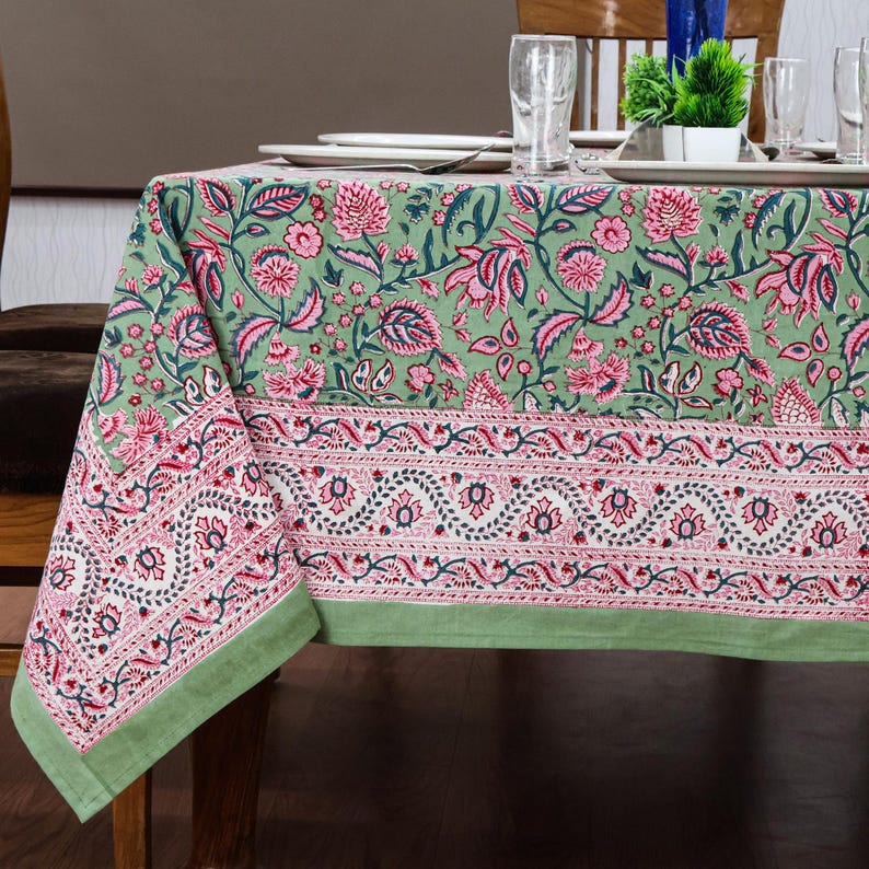 Beautiful hand block printed cotton tablecloth featuring floral patterns with pink and green accents. This vibrant Indian dining table cover adds a touch of bohemian charm to your kitchen or dining space. Ideal for everyday use, festive meals, or special occasions like housewarmings, weddings, baby showers, or dinner parties. Styled on a wooden table with white plates, cutlery, glasses, and a centerpiece flower vase.