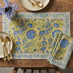 Op de afbeelding: Een rechthoekige placemat en een gevouwen servet met een gele achtergrond, een herhalend blauw bloemenpatroon en groen blad. De set bevat een rand met een blauw en wit geometrisch ontwerp dat past bij de stijl van het hoofdmotief.