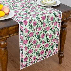Può includere: Un runner da tavola rettangolare con sfondo verde chiaro e un motivo ripetuto di rose rosa e foglie verde scuro. Il runner presenta un bordo distinto con una stampa floreale più piccola ed è posizionato su un tavolo in legno.