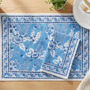 Puede incluir: Un mantel individual rectangular y una servilleta a juego doblada con fondo azul claro y un patrón de flores y hojas tropicales en azul oscuro. El conjunto incluye un borde decorativo con un motivo circular y un borde exterior blanco.