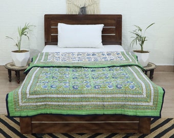 May include: A green and white floral patterned quilt with a navy blue border. The quilt is folded on a wooden bed frame with a white pillow.