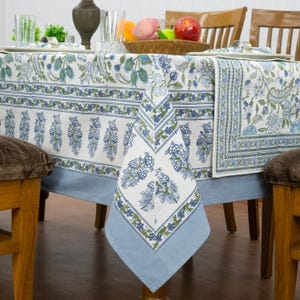 Pode incluir: Uma toalha de mesa branca com um padrão floral estampado em blocos azuis e verdes, com uma borda azul-clara sólida. O tecido cobre uma mesa de jantar, exibindo motivos botânicos complexos, incluindo flores e folhas estilizadas num design repetitivo.