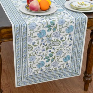 Könnte beinhalten: Ein weißer Tischläufer mit einem sich wiederholenden blau-grünen Blumenmuster, komplizierten botanischen Motiven und einer dekorativen Bordüre. Der Läufer liegt auf einem Holztisch, auf dem ein weißer Teller mit einem Pfirsich und einer Orange steht.