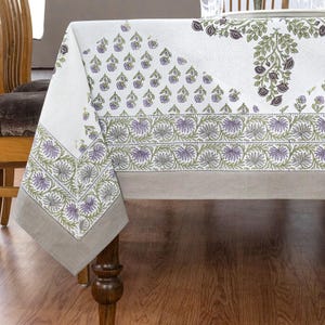 May include: A white tablecloth with a floral pattern in shades of purple and green. The design includes repeating floral motifs and a border with stylized leaves. The tablecloth has a neutral-colored border and is draped over a wooden table.