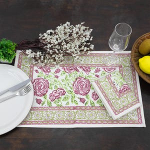 Puede incluir: Un mantel individual rectangular y una servilleta plegada con fondo blanco y un patrón repetitivo de rosas rosas, hojas verdes y un borde decorativo en rosa y verde. El conjunto se muestra con un plato blanco, cubiertos y un vaso.