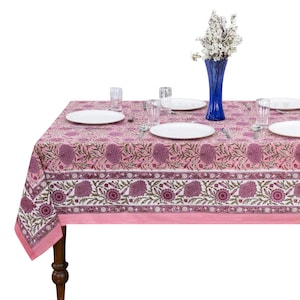 May include: A rectangular tablecloth featuring a pink and white floral block print pattern with dark purple flower motifs. The table is set with four white plates, four clear glass tumblers, and a tall blue glass vase containing white flowers.
