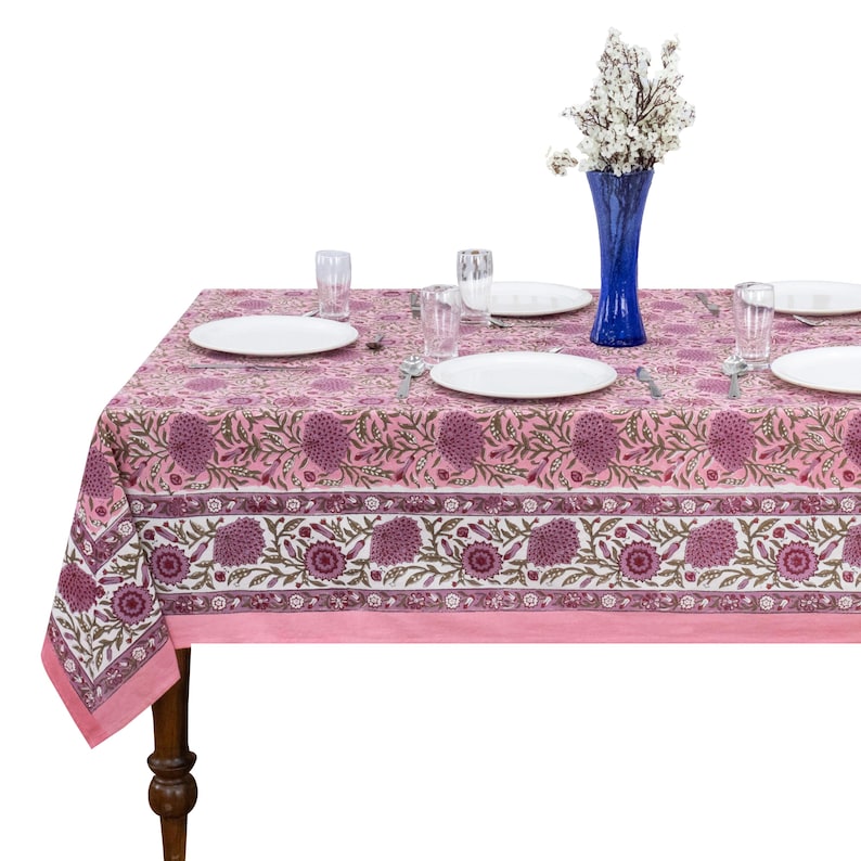 May include: A rectangular tablecloth featuring a pink and white floral block print pattern with dark purple flower motifs. The table is set with four white plates, four clear glass tumblers, and a tall blue glass vase containing white flowers.