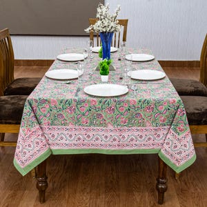 Beautiful hand block printed cotton tablecloth featuring floral patterns with pink and green accents. This vibrant Indian dining table cover adds a touch of bohemian charm to your kitchen or dining space. Ideal for everyday use, festive meals, or special occasions like housewarmings, weddings, baby showers, or dinner parties. Styled on a wooden table with white plates, cutlery, glasses, and a centerpiece flower vase.